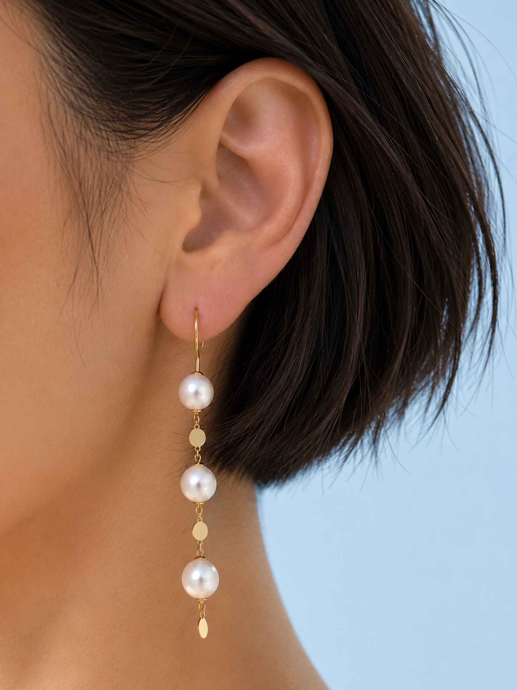 A close-up of a model's ear wearing a long gold drop earring with four white pearl beads and small gold disc accents, set against a solid light blue background.