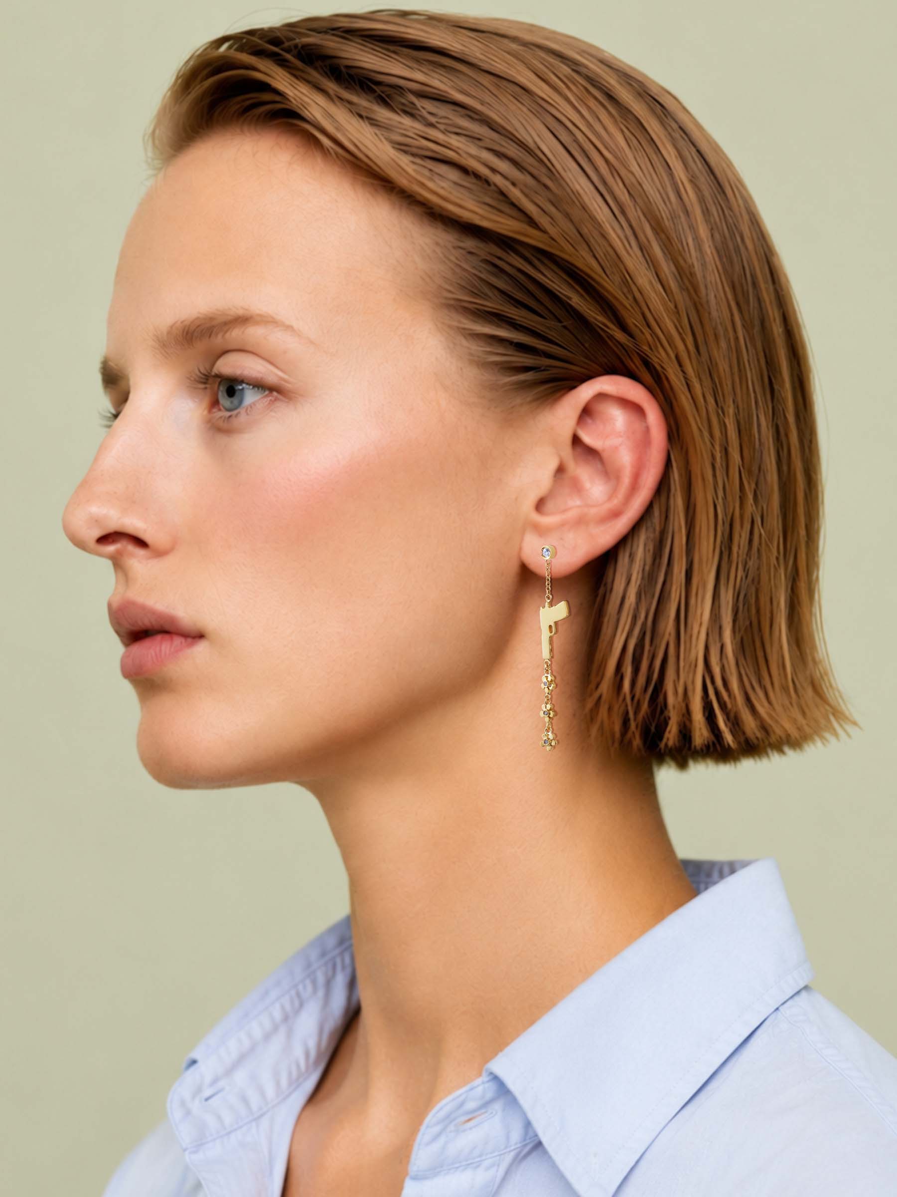 A woman in profile wearing a delicate gold dangle earring with small dangling charms, against a solid light beige background.