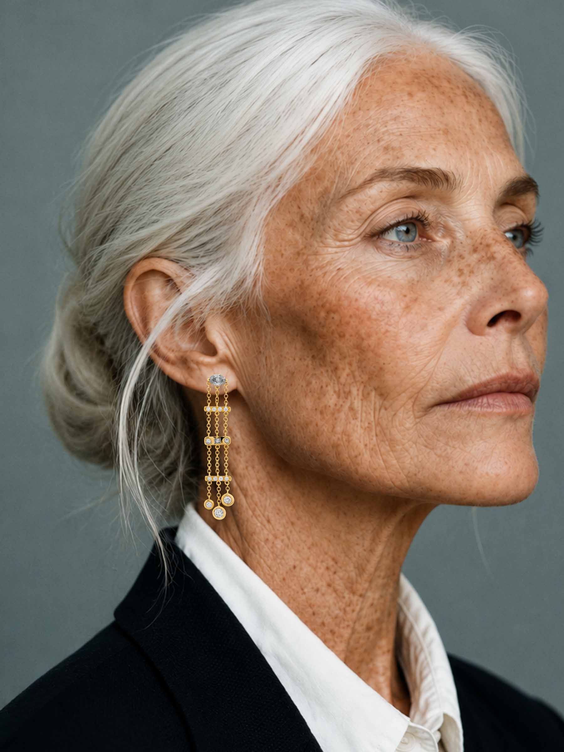 Close-up portrait of an older woman with white hair pulled back, wearing a long gold dangle earring with small clear stones, against a plain gray background.