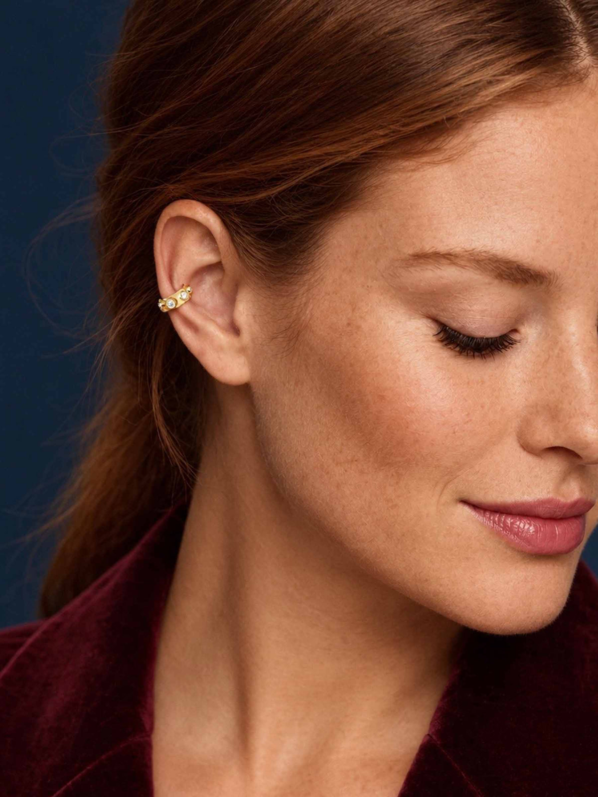 A close-up of a woman with brown hair wearing a delicate gold ear cuff with small clear stones, set against a dark blue background.