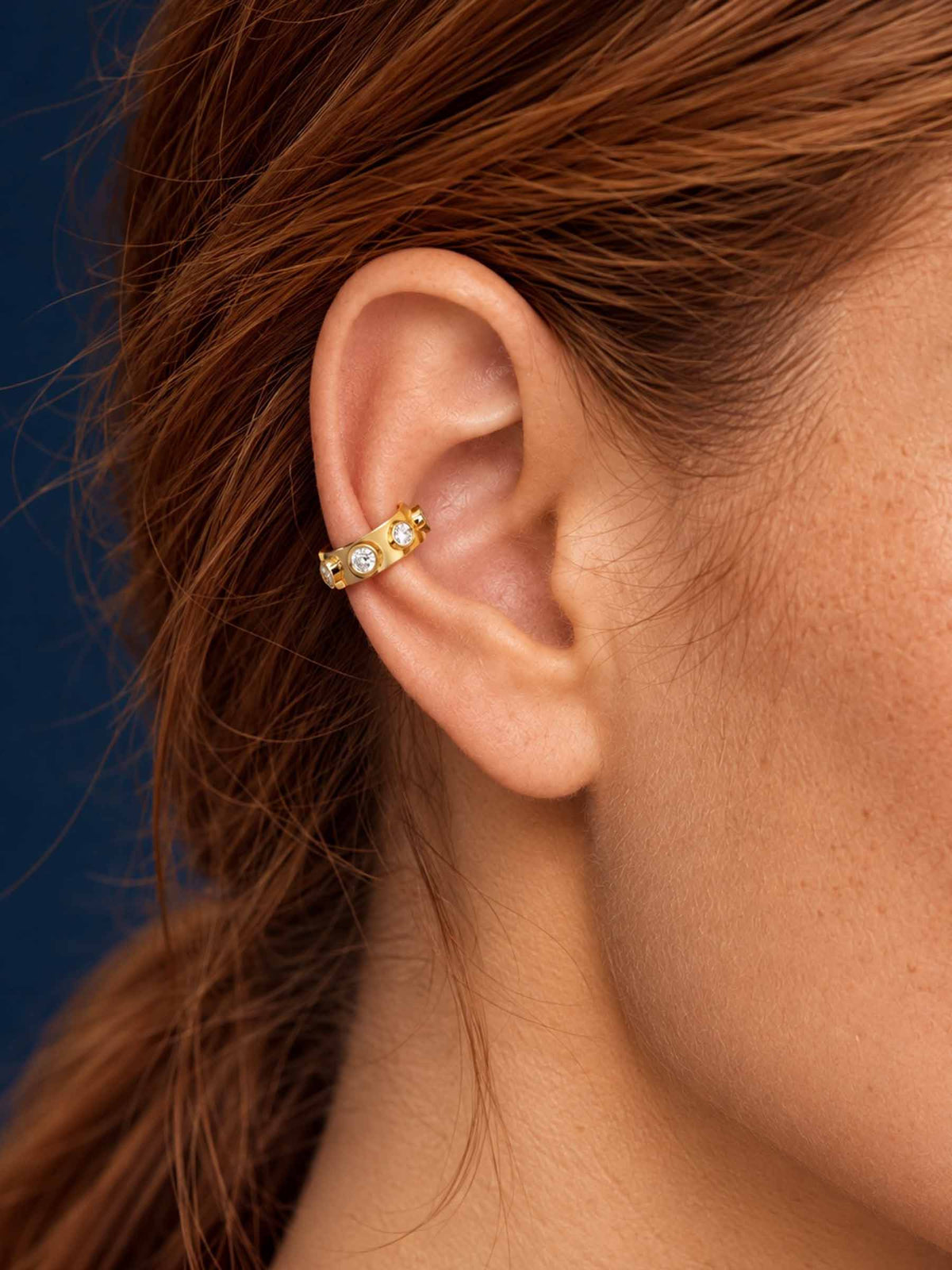 A close-up of a woman's ear wearing a delicate gold ear cuff adorned with small clear gemstones, set against a dark blue background.