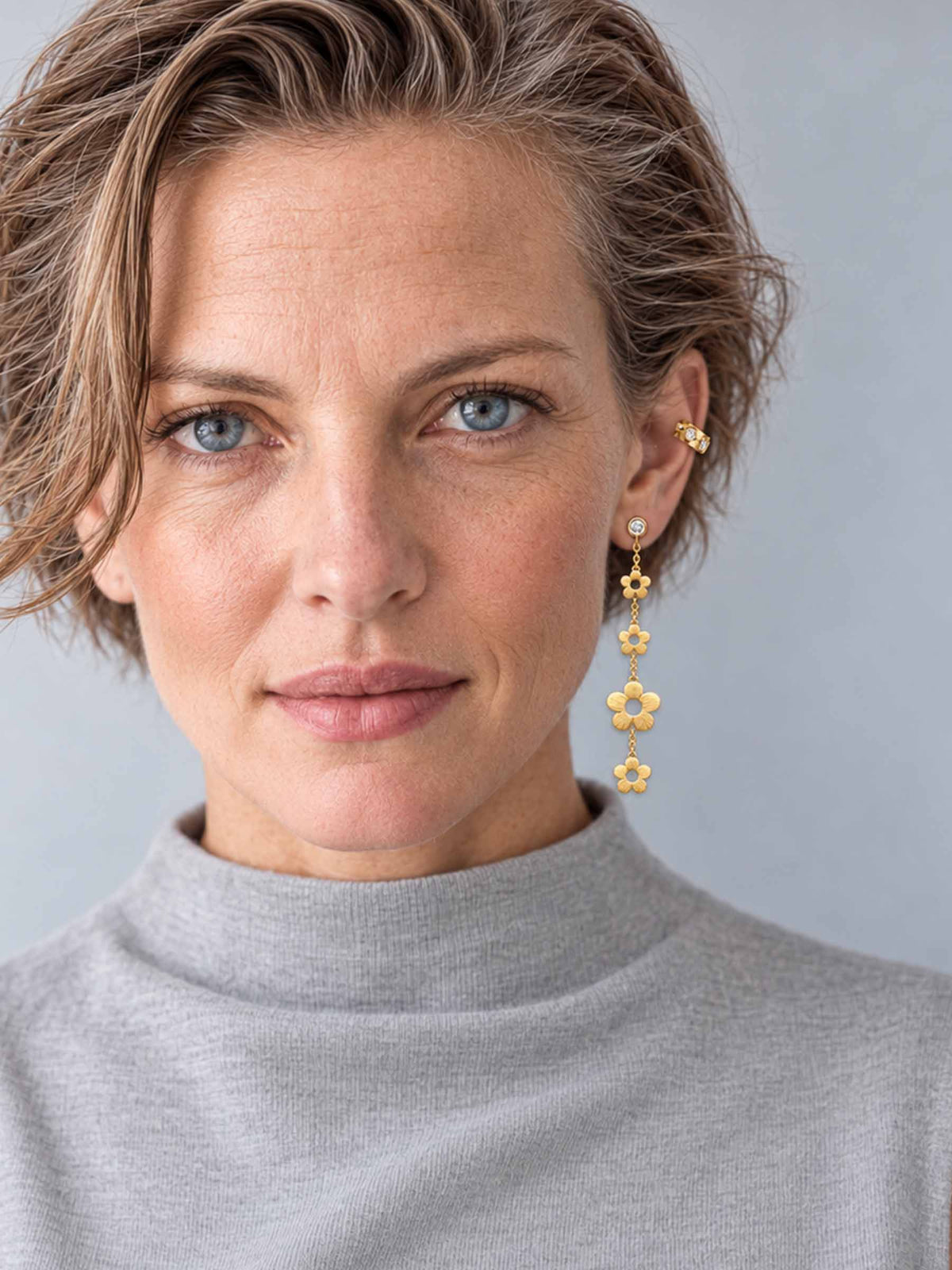 A close-up portrait of a woman with short light brown hair wearing a gray mock neck top and a long, dangling gold flower earring. The background is a solid, neutral gray.