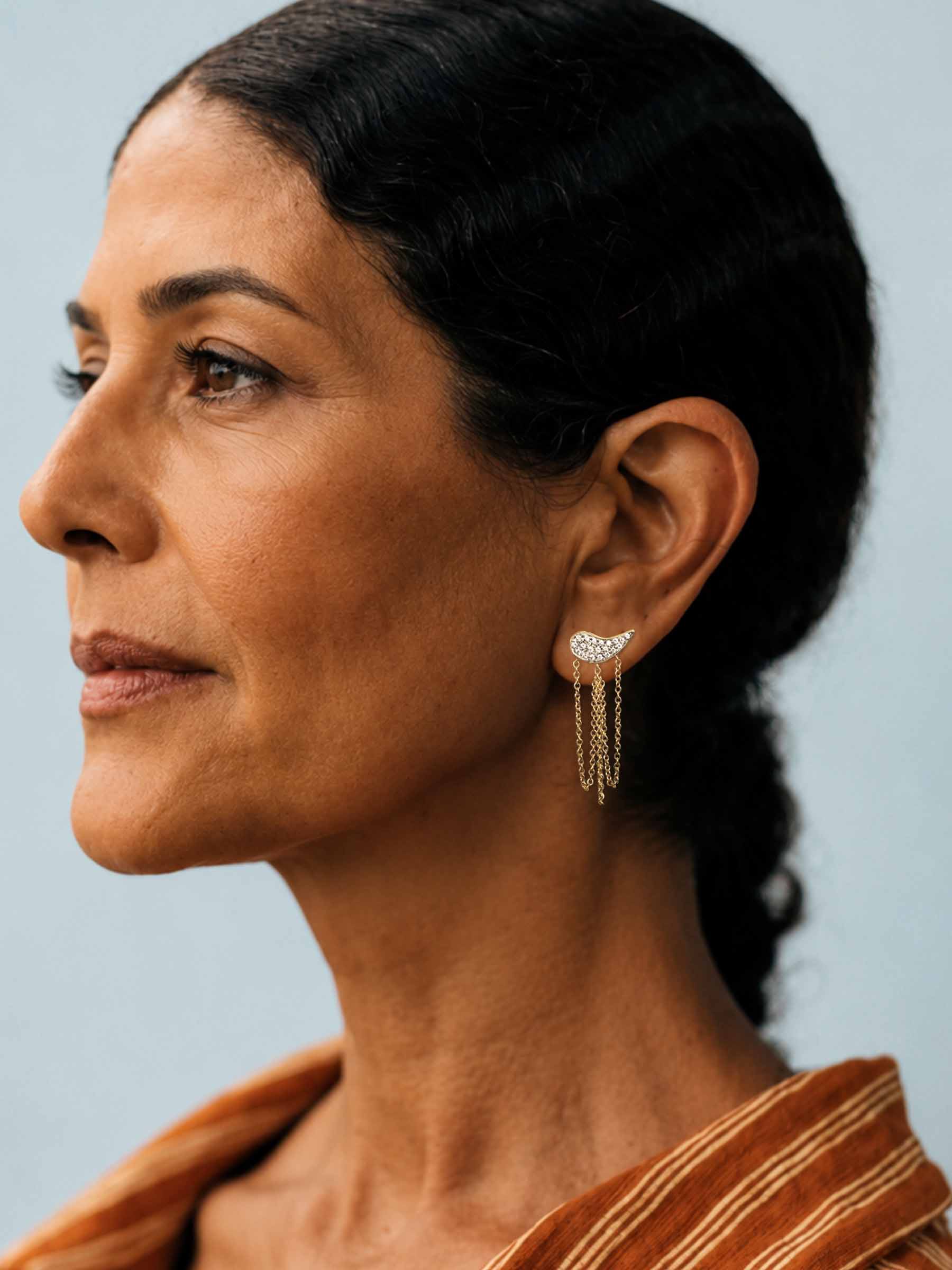 A close-up side profile of a woman with dark hair pulled back, wearing a delicate gold crescent-shaped dangle earring with small clear stones and three hanging chain drops. She is wearing a rust-orange striped top against a solid light blue-gray background.