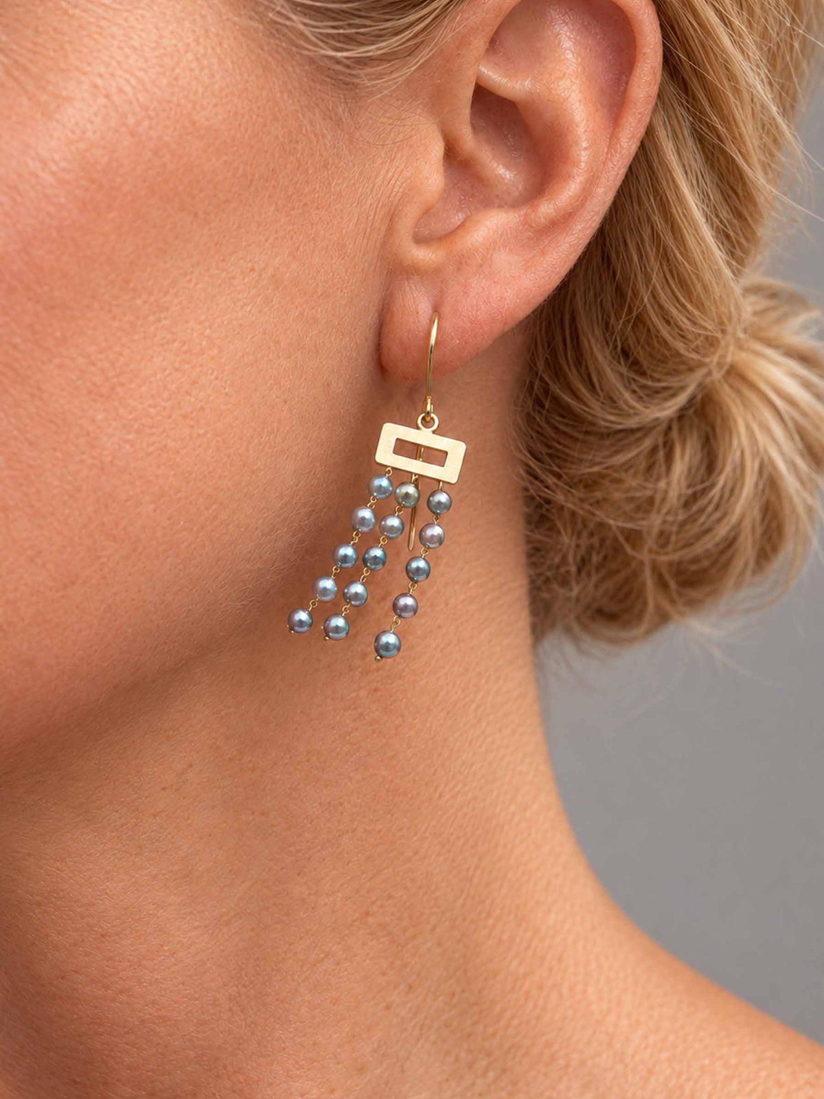 A close-up of a model's ear wearing a gold dangle earring with three strands of small gray-blue pearls.