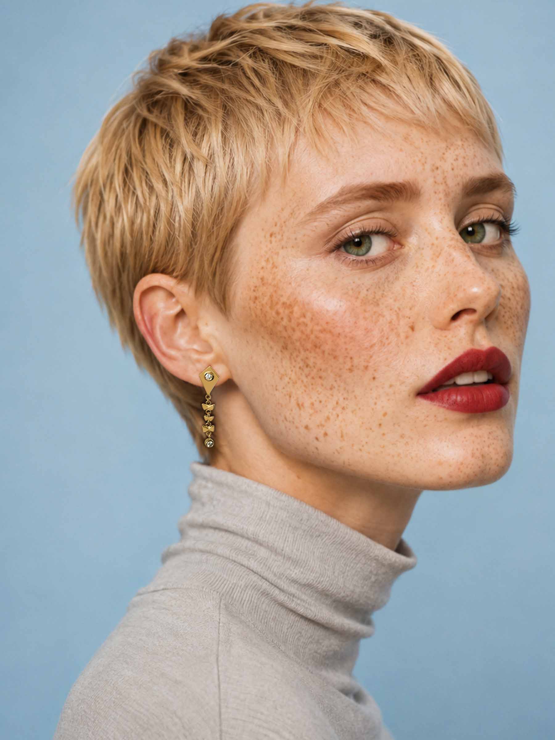 A close-up portrait of a woman with short blonde hair, freckles, and red lipstick, wearing a gray turtleneck and a gold dangling earring against a light blue background.