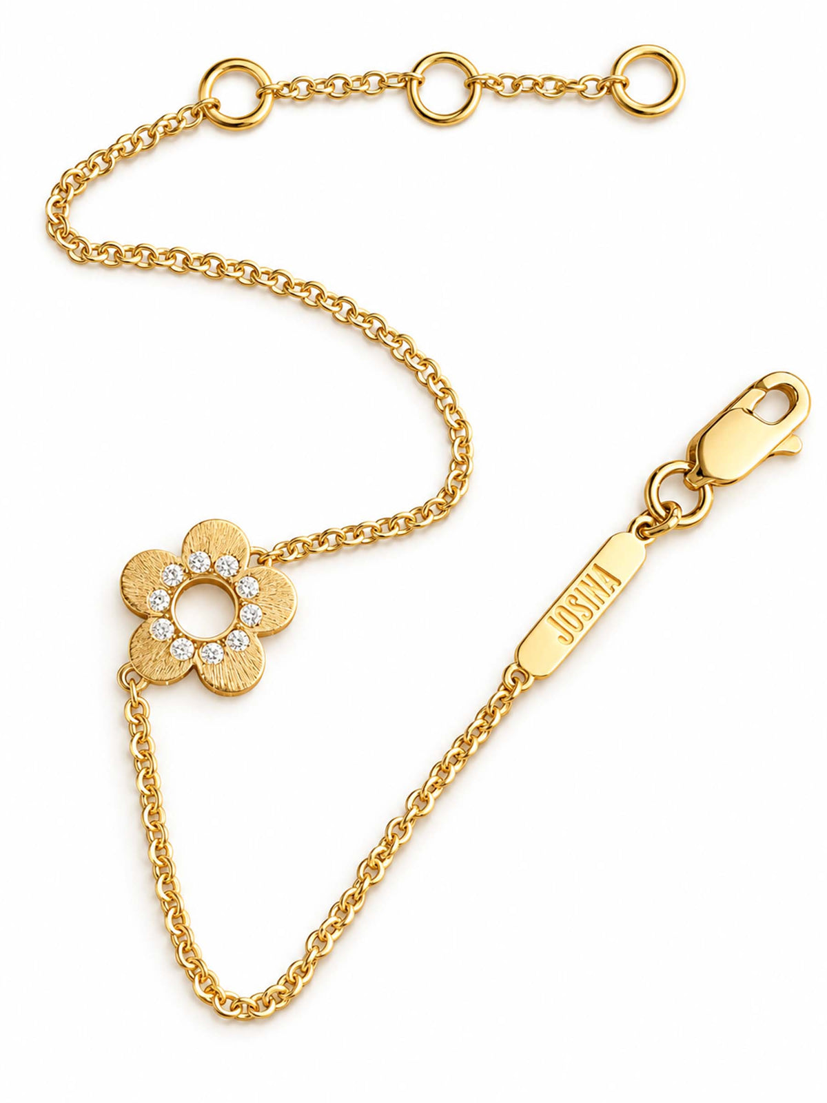 A gold chain bracelet with a flower-shaped charm set with small clear stones, shown on a plain white background. The chain includes an extender section with three rings and a lobster clasp, with a brand tag engraved with JOSINA