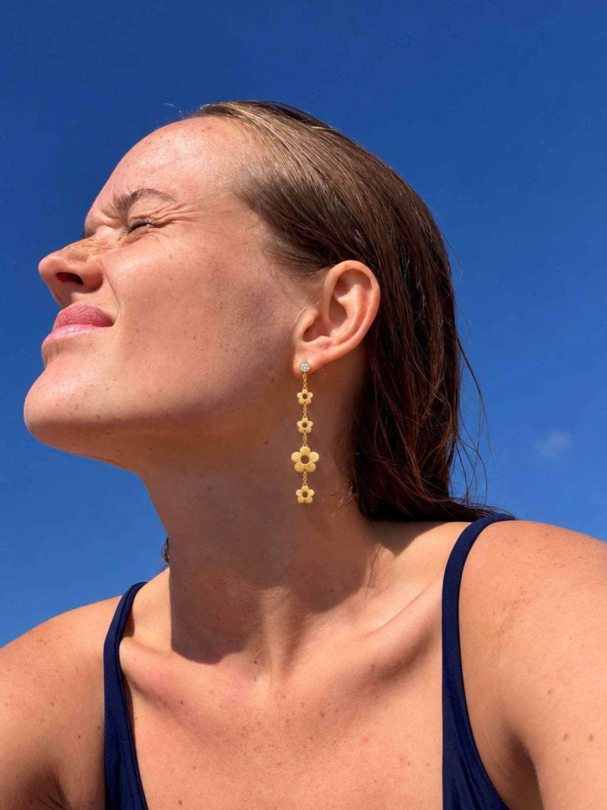 A woman outdoors under a clear blue sky wearing a long gold dangle earring with four small flower-like links.