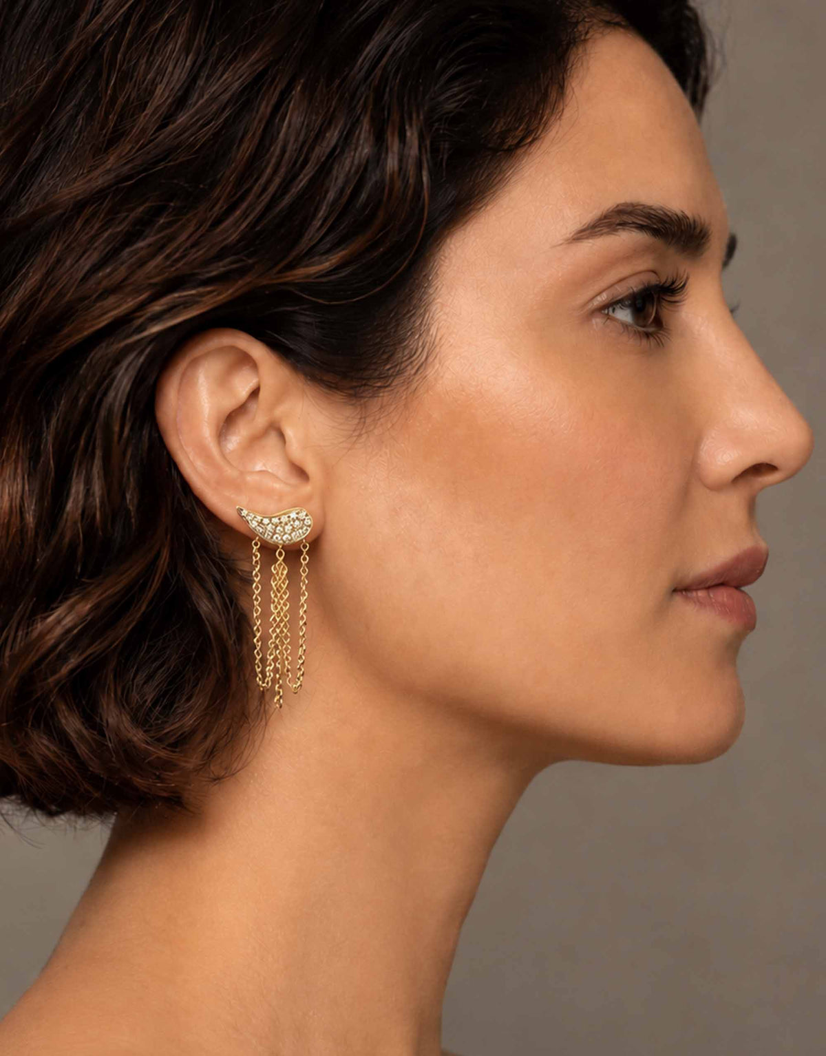 A close-up side profile of a woman wearing a delicate gold dangle earring with a small cluster of clear stones at the top and three hanging chain drops.
