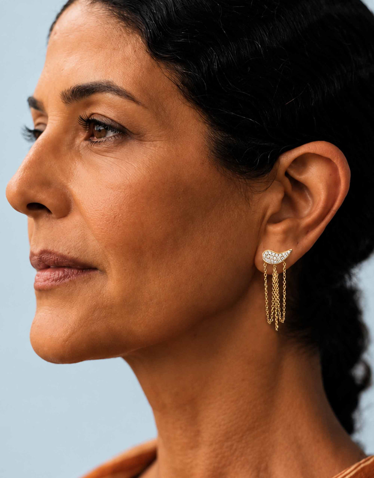 A close-up side profile of a woman wearing a gold crescent-shaped dangle earring with small clear stones and three dangling chain strands against a light blue background.