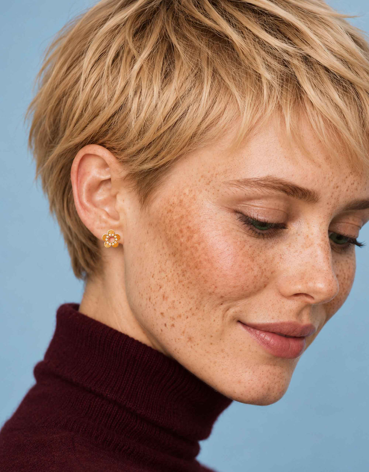 A close-up of a woman with a short blonde pixie cut and freckles, wearing a small gold flower-shaped stud earring, against a solid light blue background.