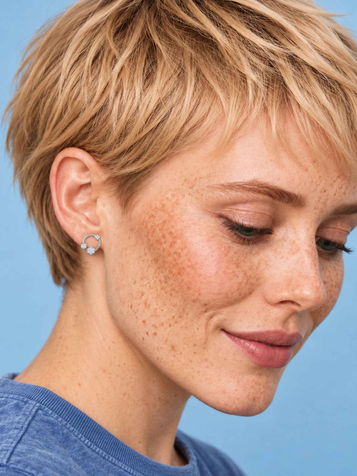 A close-up of a woman with short blonde hair and freckles wearing a small silver hoop earring with a clear gemstone, set against a solid blue background.