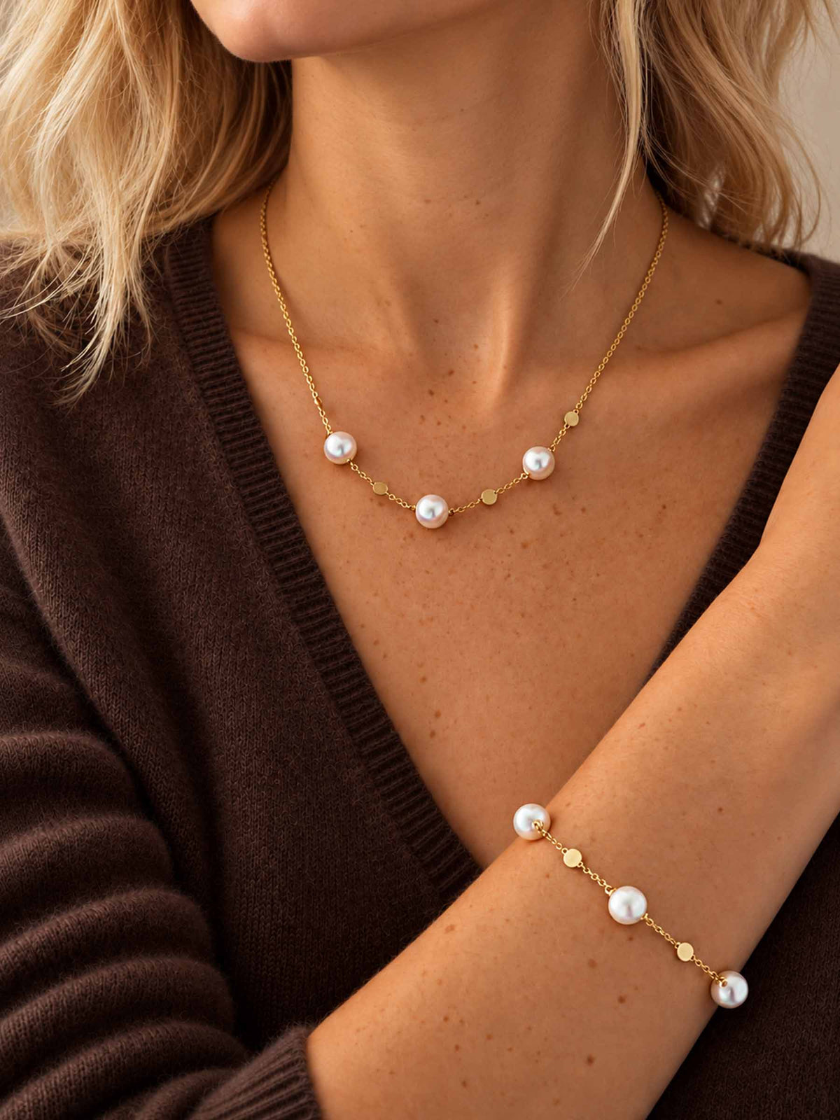 A close-up lifestyle shot of a model wearing a delicate gold necklace and matching bracelet, both featuring white freshwater pearls and small gold dots, styled with a dark brown knit cardigan.